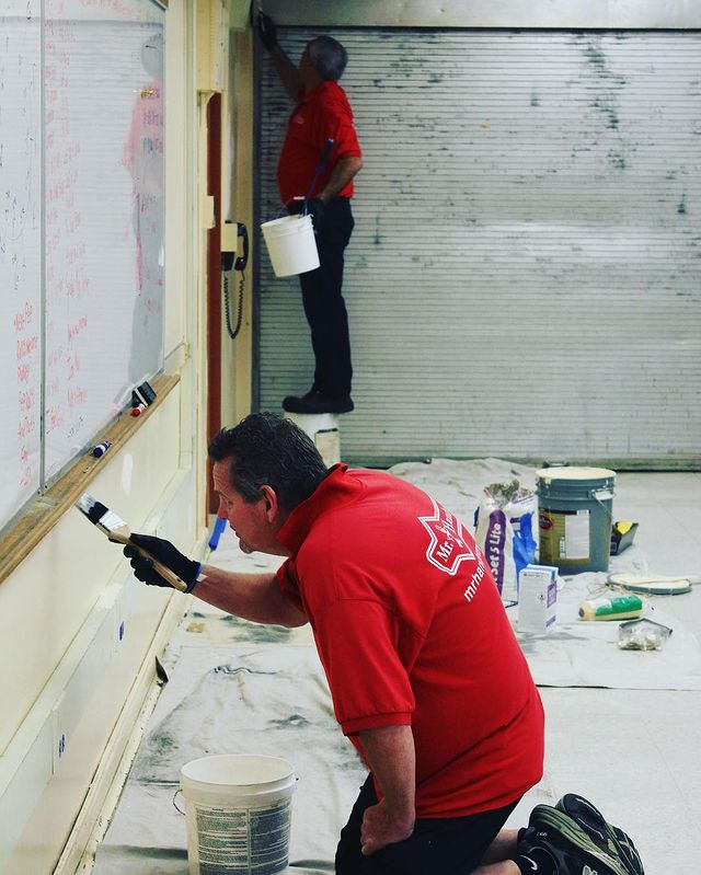 Man on knees painting a wall inside a school.