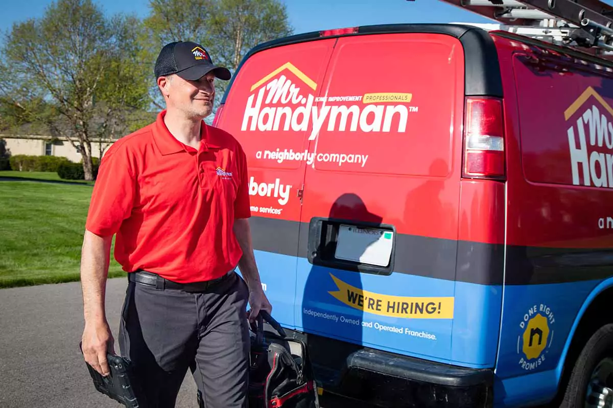 A Mr. Handyman service professional arrives at a job site in a branded van with tools in hand, ready to install a fence.