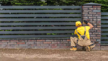 A man in a yellow shirt and cap and khaki overalls installs a metal fence