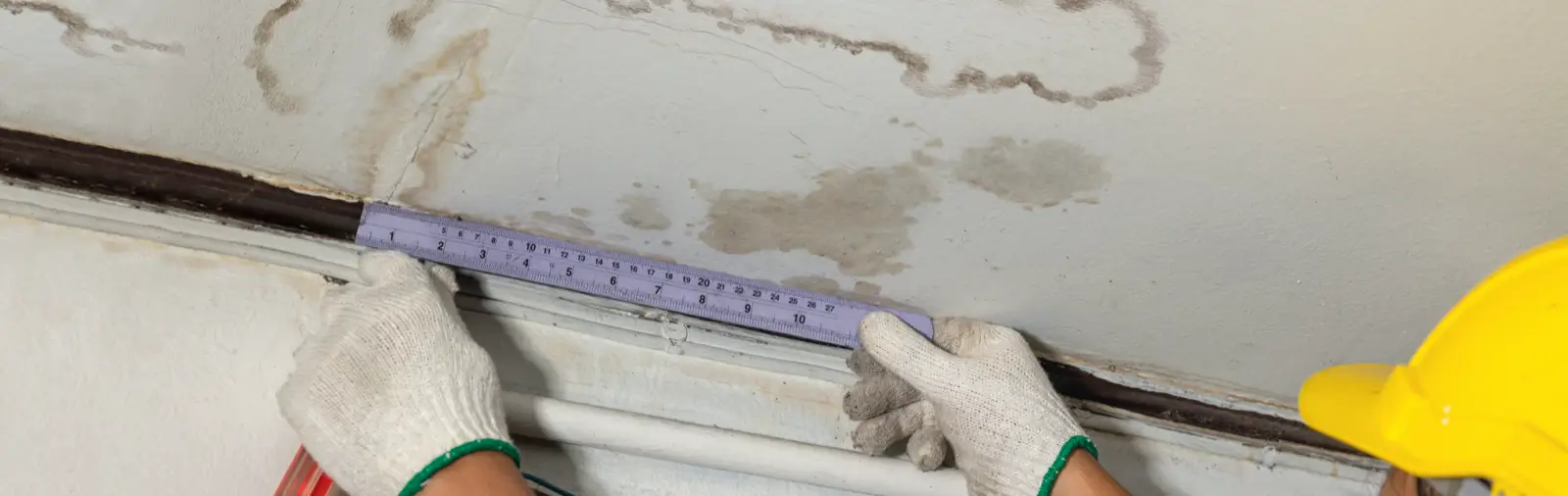 Person wearing gloves and a yellow hard hat, using a ruler to measure a water stain on a white ceiling, highlighting ceiling damage.