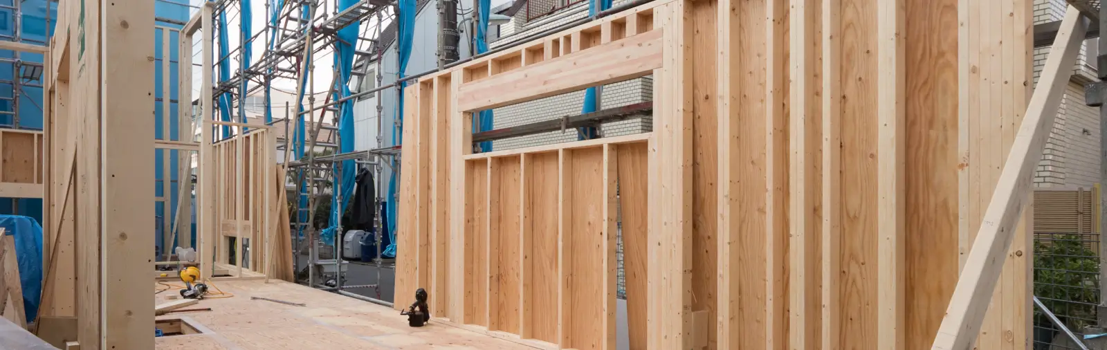 Newly constructed wooden wall frame with vertical and horizontal beams, under clear skies with building scaffolding in the background.