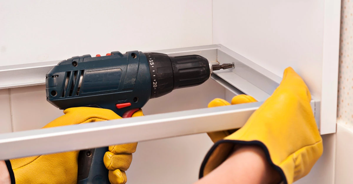 A close-up image of a handyman using a power drill to mount cabinet boxes on a wall during a cabinet installation project.