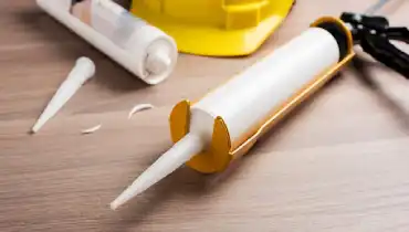 A caulking gun with a fresh tube of caulk and a used tube laying near a hard hat on the wood floor of a home.