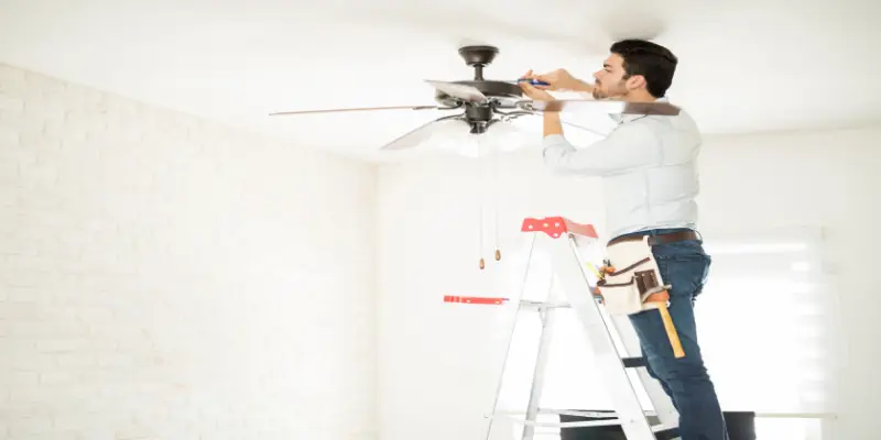 A handyman with electrical skills and a tool-belt stands on a ladder while fixing a ceiling fan