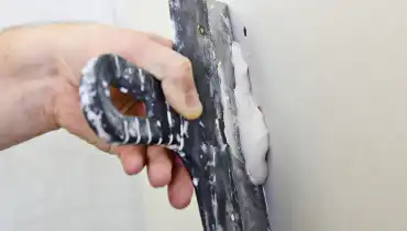 A handyman using a wide putty knife to spread fresh drywall compound across a recently completed drywall repair job.