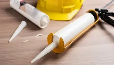 A tube of caulk loaded in a caulking gun and another empty, used tube laying next to a hardhat nearby.