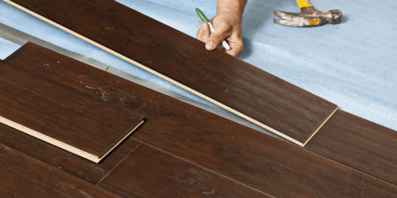 A handyman installing flooring planks across a residential subfloor with a hammer laying nearby.