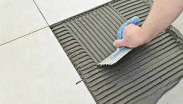 A handyman using a notched trowel to smooth out mortar in an empty space on a tile floor before installing a replacement tile during an appointment for tile repair in Vancouver, WA.