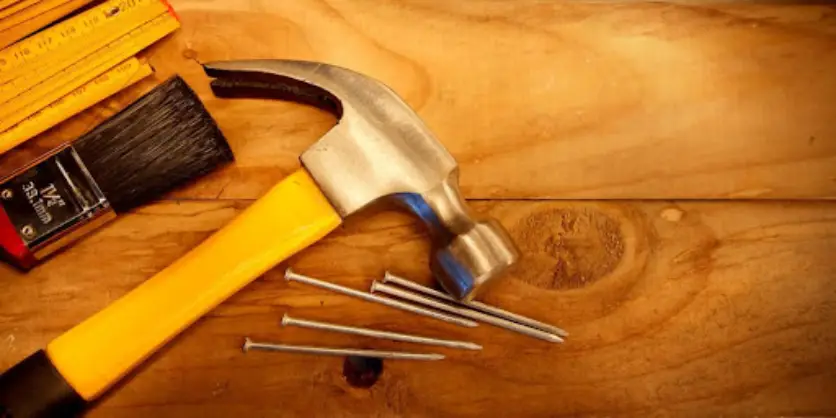 A hammer, rulers, a paintbrush, and several nails all lying on top of wooden boards.