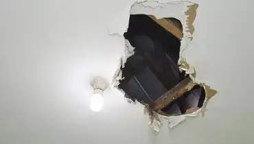 A hole in a residential ceiling located near a light fixture, with wooden ceiling beams visible in the space beyond the hole.