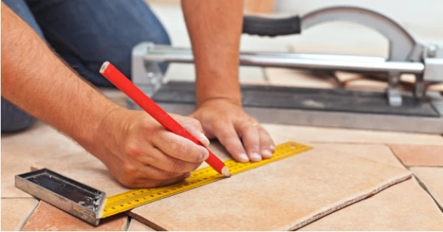 A person using measuring tape and pencil to mark a line on a loose piece of tile on the ground.