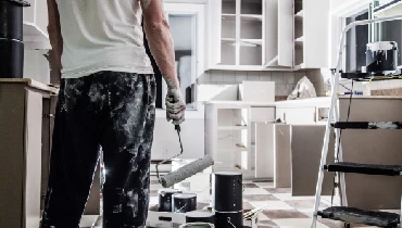 Man holding paint roller standing in kitchen surrounded by paint cans and supplies.