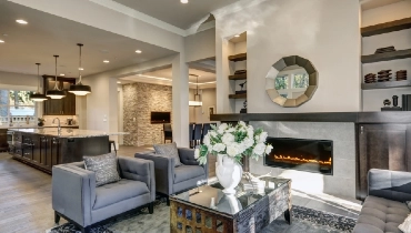 Chic living room filled with built-in cabinets flanking round mirror atop grey tile fireplace, tufted sofa facing two armchairs and window wall overlooking lush outdoors.