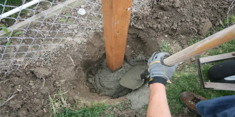 A handyman shoveling cement around a wooden fence post