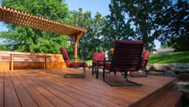 The sun shines on a deck with a fresh coat of stain, a pergola, and chairs.