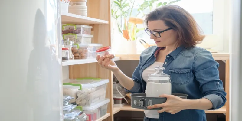 Interior of wooden pantry with products for cooking; adult woman taking kitchenware and food from the shelves.