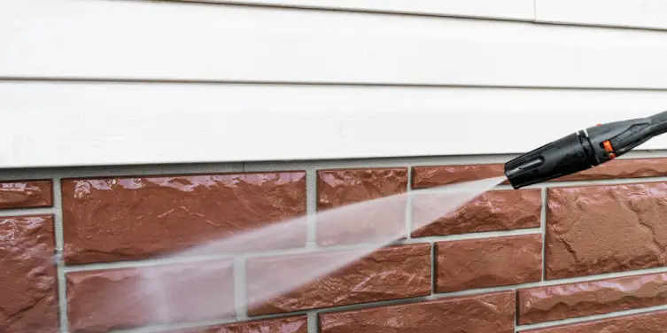 The nozzle of a power washing machine spraying jets of water on the brick siding of a home.