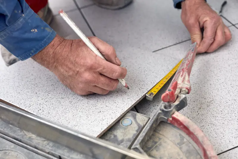 Person leaning over and measuring tile with pencil and measuring tape.