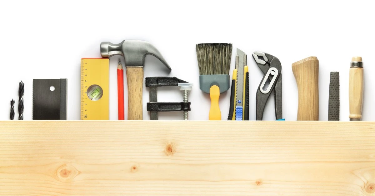 A series of tools that might be used by a handyman in Primrose, TX laid out underneath a wooden board, including a hammer, level, pencil, brush, and utility knife.