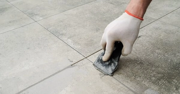 A handyman using a putty knife to apply new grout to the cracks between floor tiles after completing tile repairs in Walpole, MA.