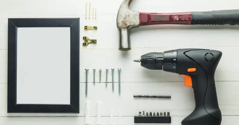 A hammer, power drill, screws, hangers, and a picture frame lying on a wood surface before being used for picture hanging service in frisco, TX.