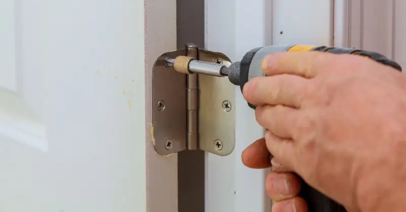 A hand holding a power drill as it is used to screw in a door hinge for a door installation project.