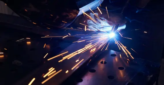 A close-up of a welder in Charleston, SC wearing a welding mask and bent over as sparks fly from a piece of metal they are working on.