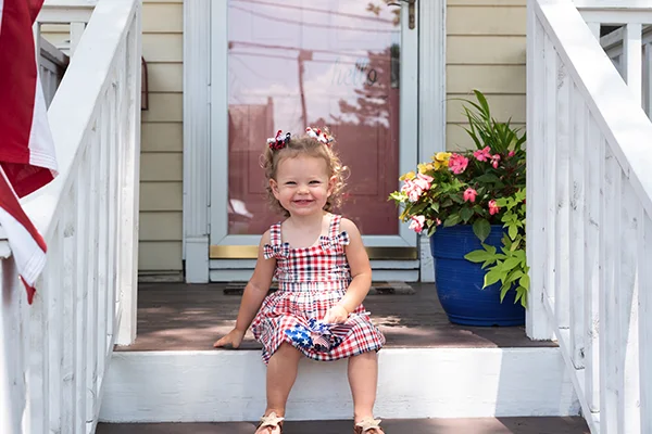 A toddler girl seated on the front porch, with a storm door visible in the background.